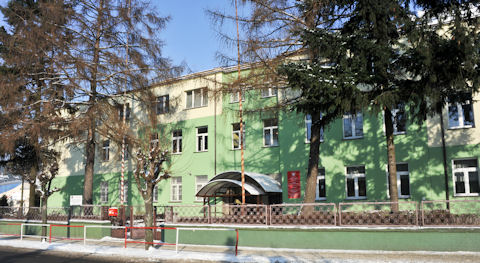Fotografia przedstawia budynek szkoły po remoncie w 2008 roku. Elewacja jest koloru zielonego z białą stolarką okienną. Przed budynkiem widać drzewa iglaste znacznie wyższe od szkoły.